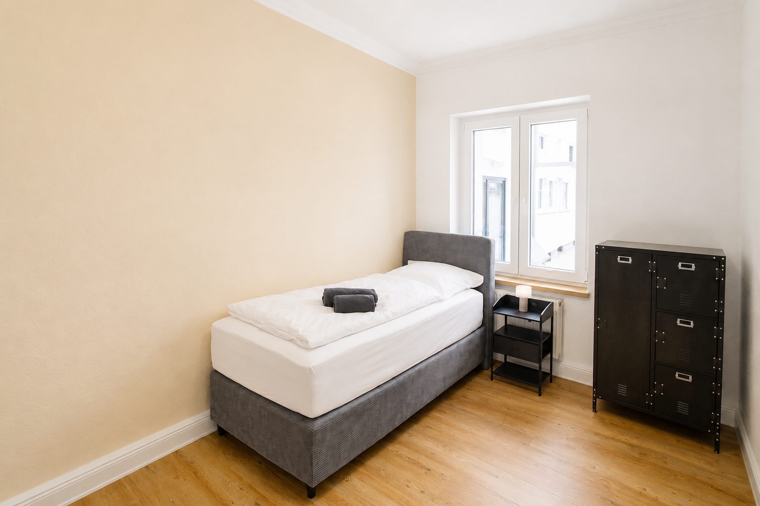 Zimmer 2 mit Einzelbett, schwarzem Metallschrank und Nachttisch mit Lampe